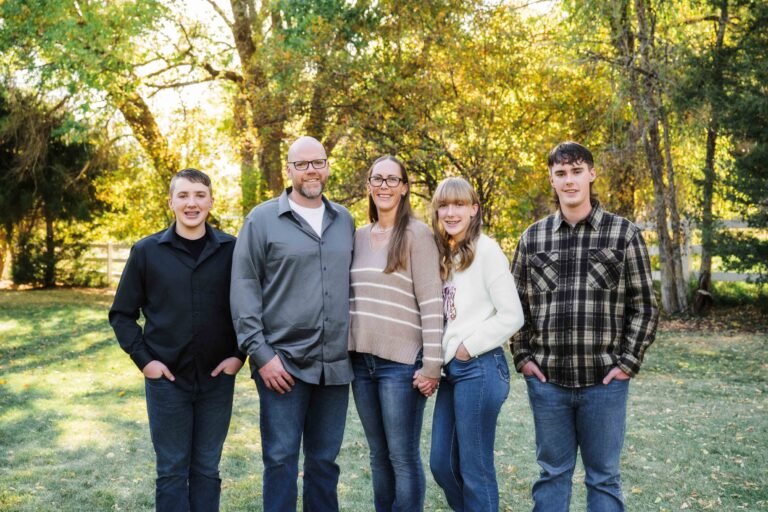 family of 5 standing on a grassy lawn with a white fence and trees behind them