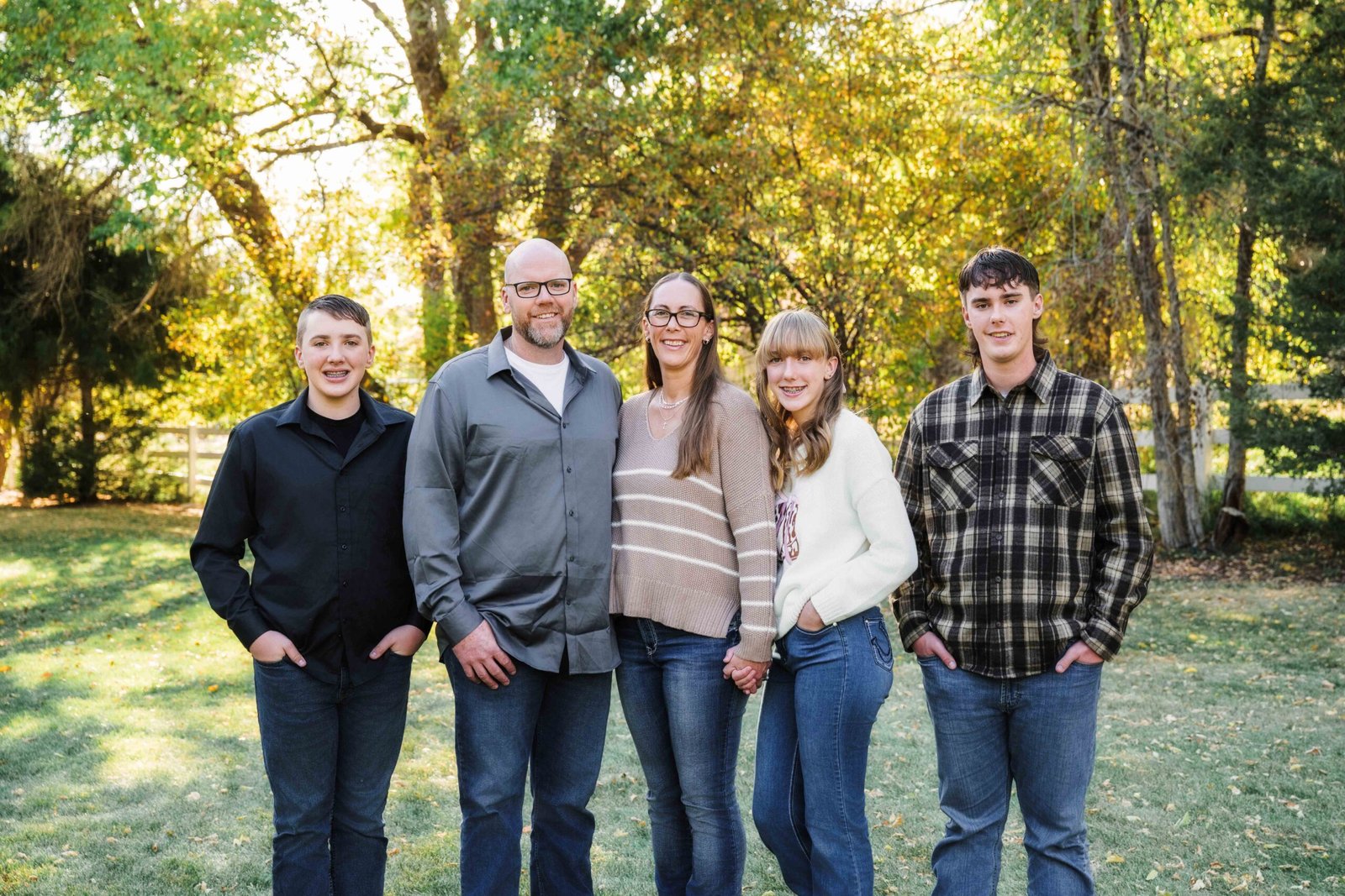 family of 5 standing on a grassy lawn with a white fence and trees behind them