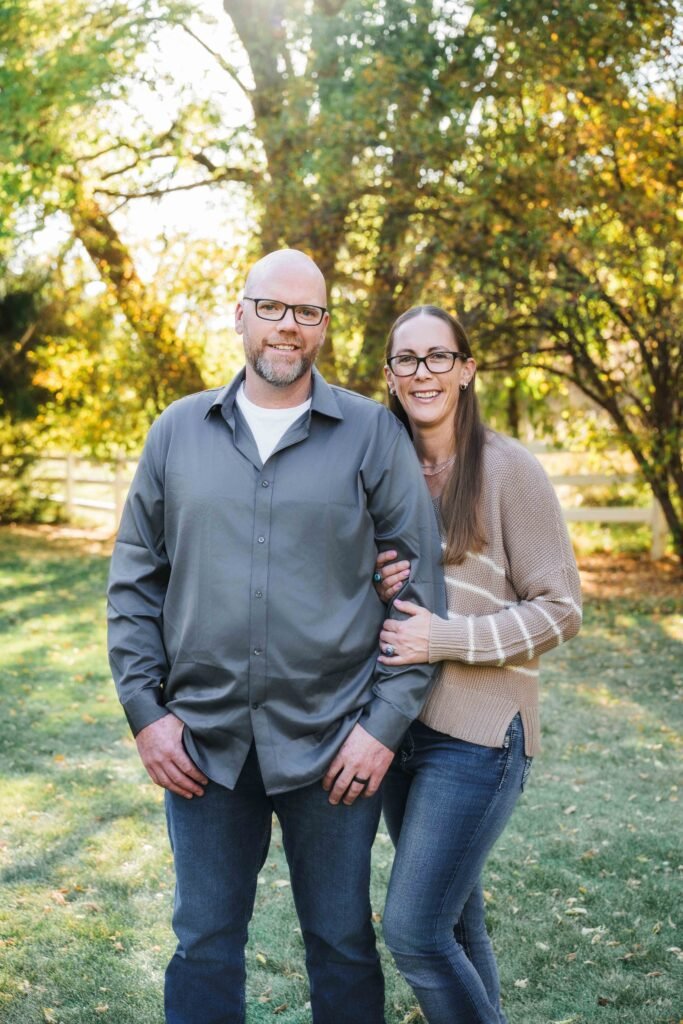 dad and mom standing for a picture, grassy lawn, white fence and trees behind them