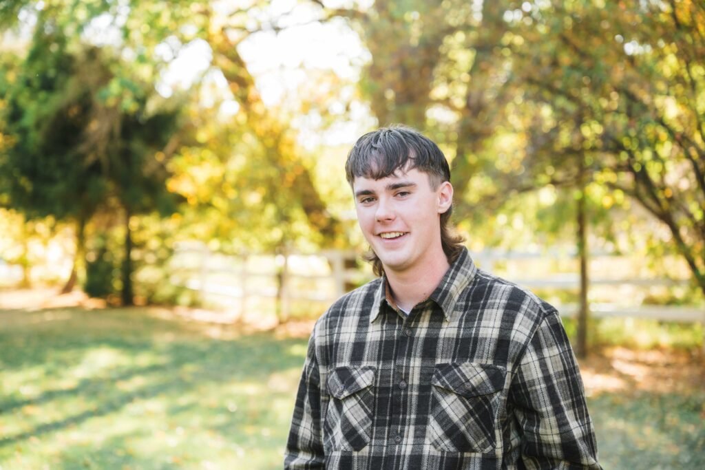 teenage boy standing for a picture, grassy lawn, white fence and trees behind him